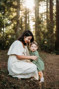 pregnant mama crouches down near her toddler daughter with redwoods behind them