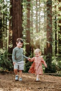 little boy holds his baby sisters hand in a forest