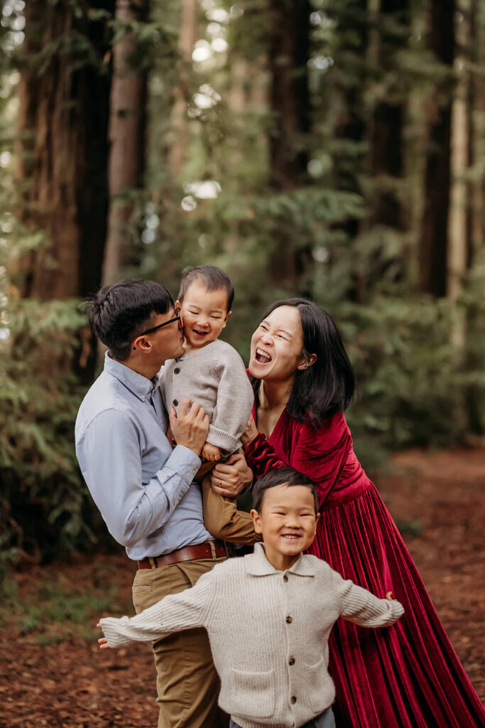 Family laughing together in a redwood forest, with parents holding their toddler while their older child runs joyfully toward the camera.