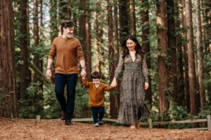 little boy walks while holding his parents hands. oaklands redwoods are behind them