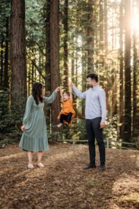 toddler girl jumps while holding hands with her parents. the sun is peaking through redwood trees behind them.