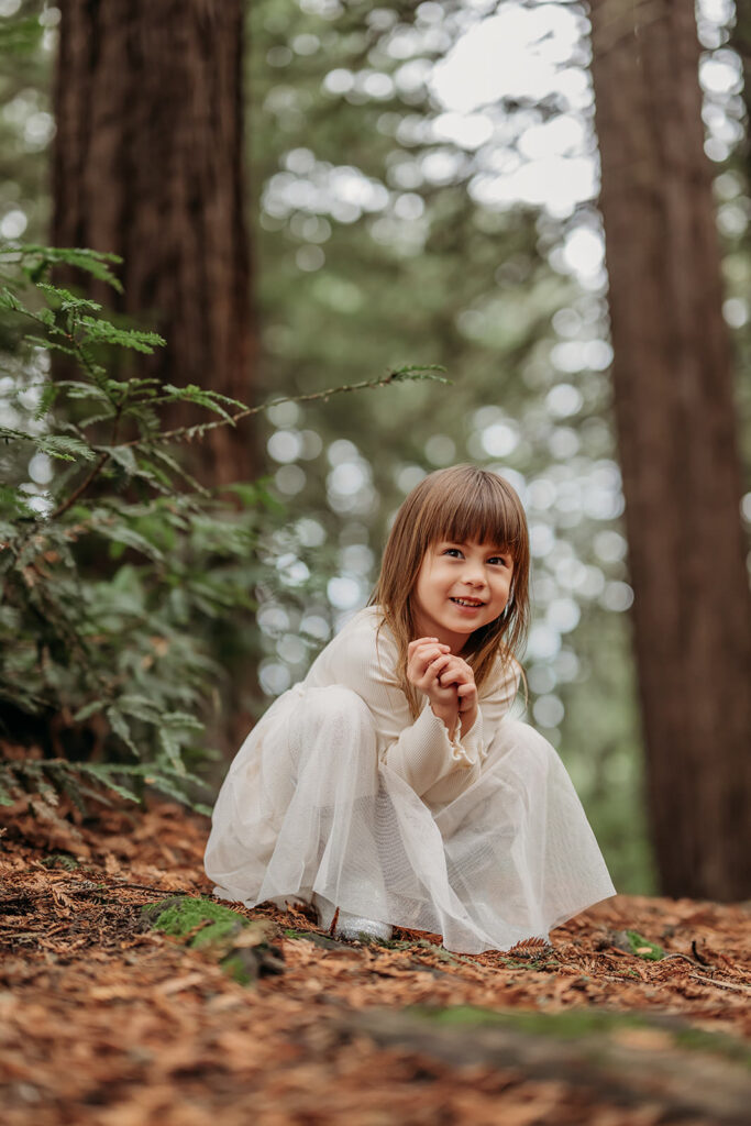 Young child sitting on the forest floor among redwood trees, wearing a light dress and smiling during an outdoor family photo session.