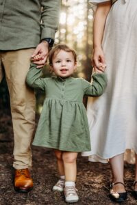 a toddler holds her parents hands and smiles at camera
