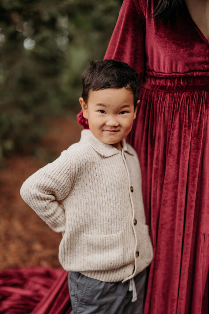 Young child standing confidently beside a parent in a forest setting, wearing a cozy sweater during a candid family photography session in walnut creek