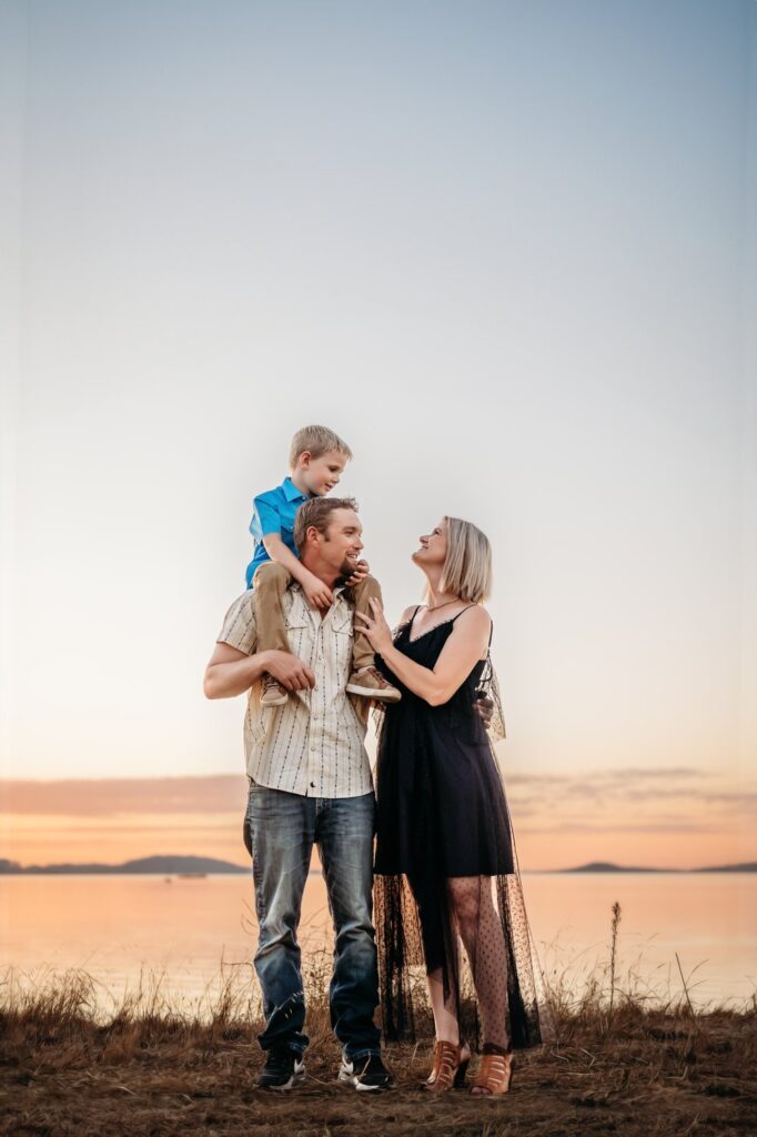 Little boy riding on dad’s shoulders while mom smiles beside them at sunset.