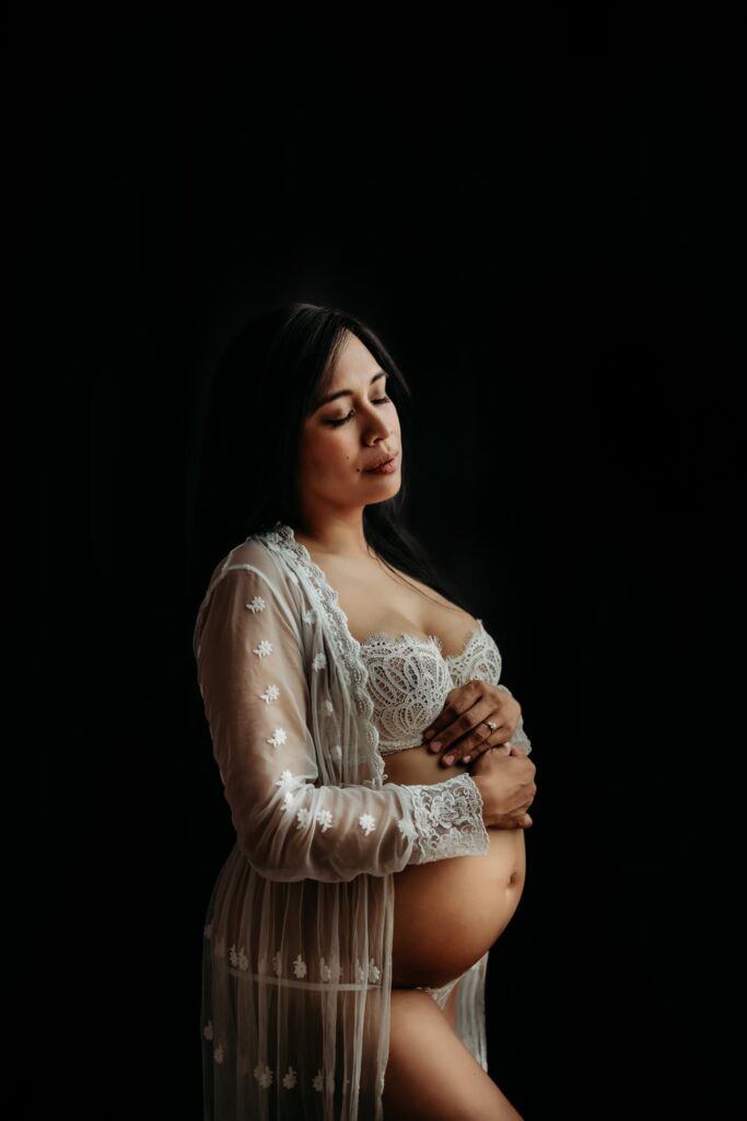 simple maternity photo with a mom in a white lace outfit on a solid black studio backdrop