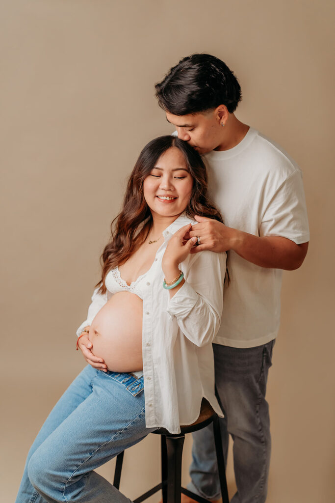 pregnant mama on her husband in a simple studio maternity session. she wears jeans and an open button down shirt