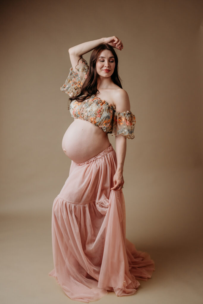 elegant studio photo of a pregnant mama wearing a 2 piece maternity gown. she poses on a minimalistic tan backdrop