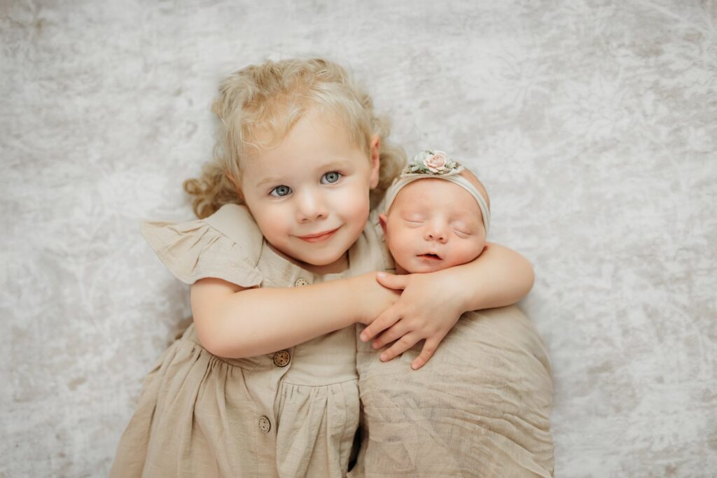 big sister hugs her baby sister while looking up at the camera during their photography session
