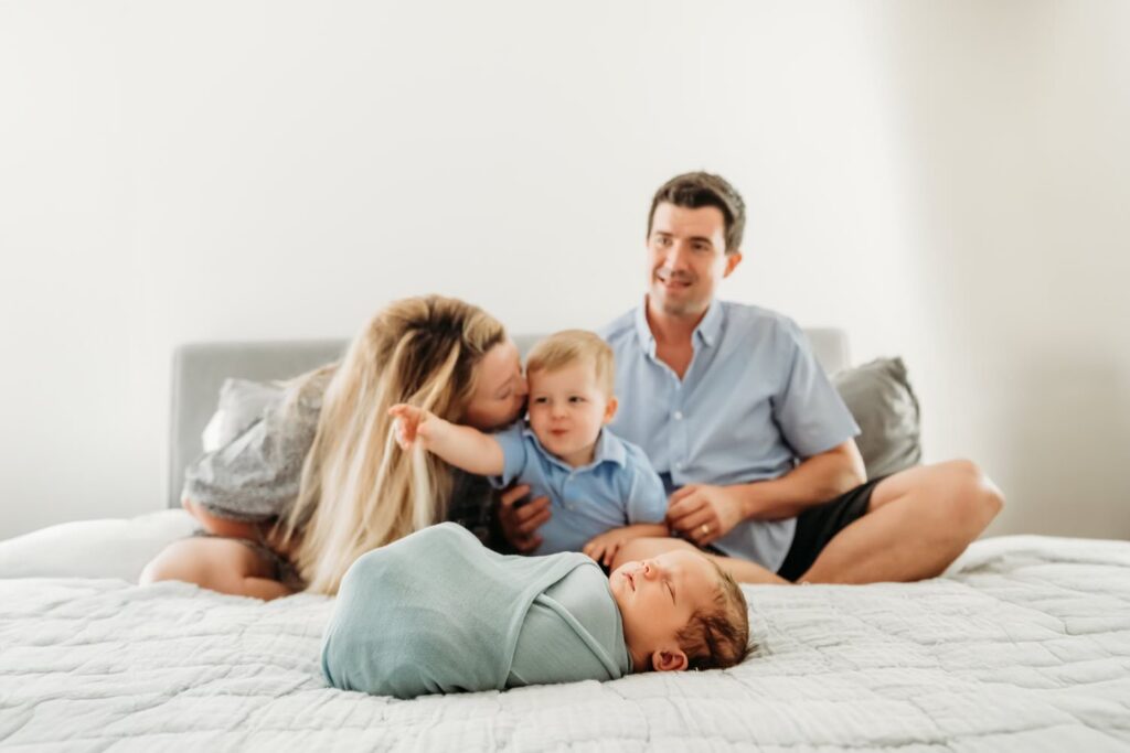 newborn baby on the foot of the bed while mom dad and big brother sit behind him