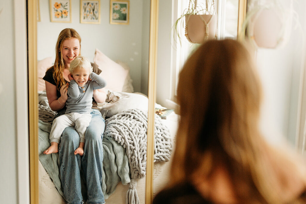 toddler sits on his sisters lap and the look at each other in the mirror