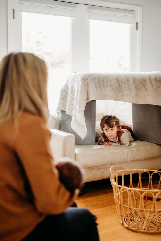 toddler hides in a pillow fort and looks over at his mom holding the newborn baby