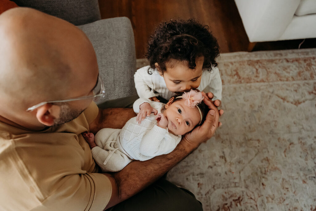 dad holds a newborn baby while their toddler kisses her head.