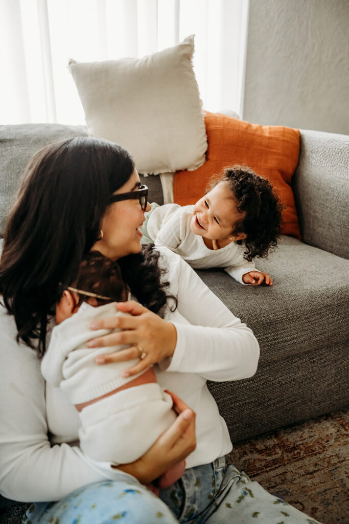 A toddler laughs on a bed while a mom holds her newborn during an in-home family photography session.